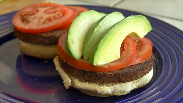 A vegan chef cooking and assembling two healthy tomato and avocado vegetarian burgers on a blue plat