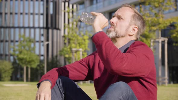 A Middleaged Handsome Caucasian Man Drinks Water From a Plastic Bottle As He Sits in a Street