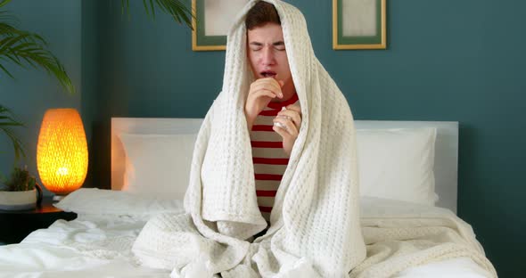 Young ill man sneezing in tissue blowing runny nose sitting at home covered with a blanket.