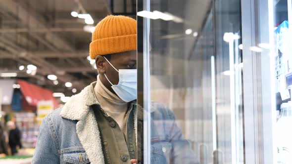 Young AfricanAmerican Man in a Medical Mask is Shopping in a Supermarket Goes to the Refrigerator