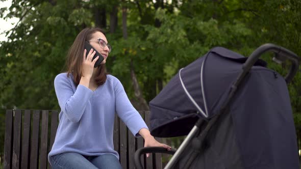 A Young Mother Speaks on the Phone and Shakes a Stroller with a Child in the Park