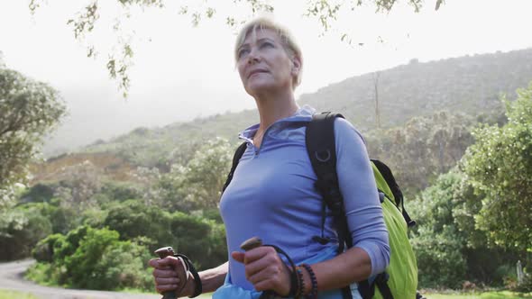 Senior hiking woman with bag pack and hiking poles standing and looking around in the woods