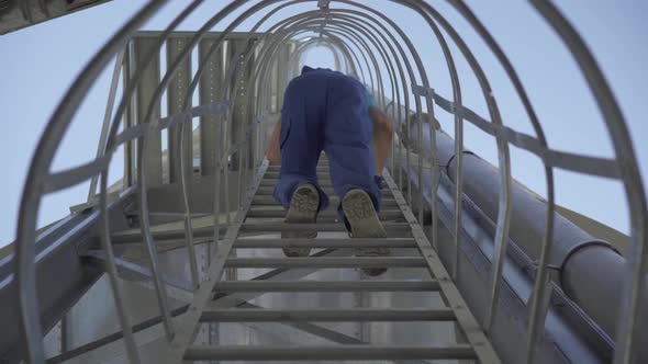 Bottom View of Factory Worker in Blue Uniform Climbing Down the Ladder on Huge Storage Tower