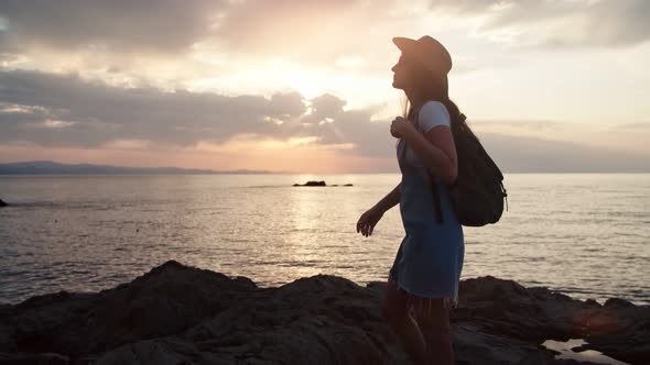 Silhouette of Female Tourist with Backpack and Hat Admiring Amazing Seascape at Sunset