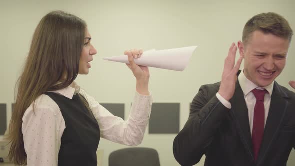 Nervous Young Girl in Formal Wear Folded Paper in the Form of a Horn and Yelling at Male Colleague