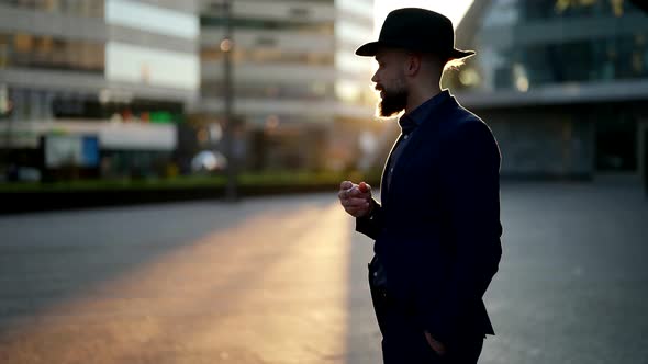 a Bearded and Mustachioed Man in a Hat Stands with a Cigarette in Hand on a Blurred City Street