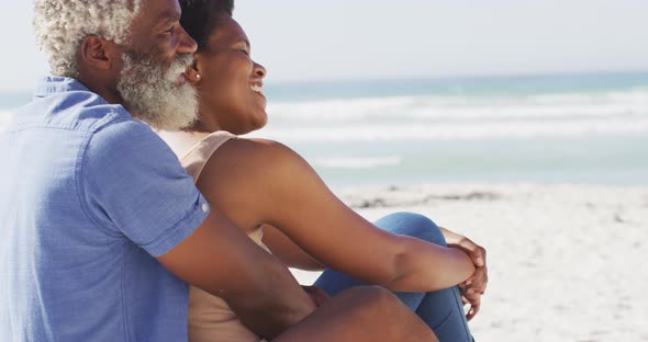 Happy african american couple sitting and embracing on sunny beach