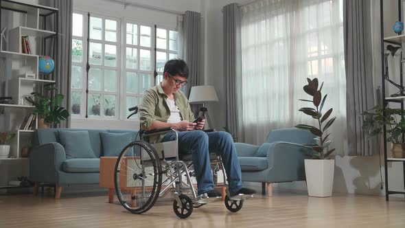 Asian Man Sitting In A Wheelchair While Smiling And Using Smart Phone At Home