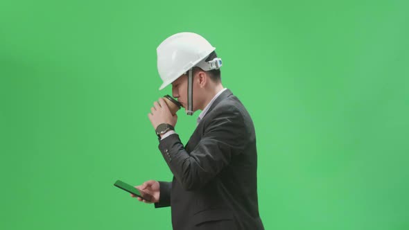 Asian Chief Engineer In The Hard Hat Use Mobile Phone And Drink Coffee While Walking On Green Screen