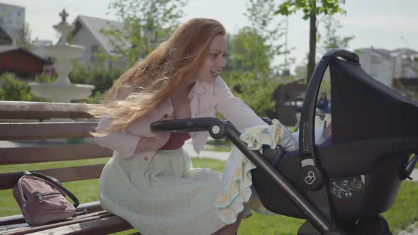 Young Happy Redhead Mother Sitting on Bench Near the House with Baby Carriage and Smiling and