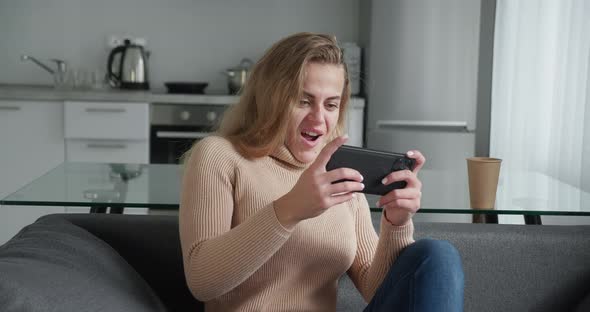 Woman in a Beige Woolen Sweater Play Game with Smart Phone on Modern Kitchen Background