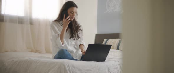 A woman talking to someone on the phone while working with a laptop from home. 