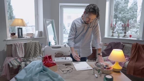 Young Creative Man Drawing with Pencil on Paper Standing at Table in Atelier