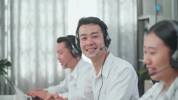 A Man Of Three Asian Call Centre Agents Smile To Camera While His Colleagues Speaking To Customer