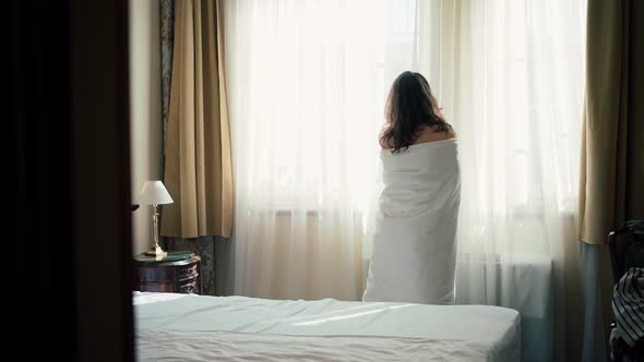 A Young Woman Wrapped in a Blanket Opens the Curtain on the Window