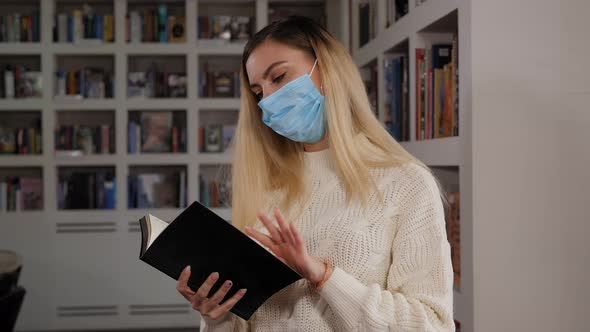 A Female Student in a Medical Mask on Her Face Reads a Book in the Library