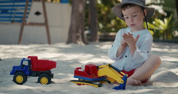 Cute Baby Boy Sitting Alone on the Beach Plays with a Plastic Crane and a Car on the Sand on the