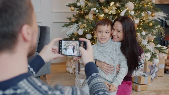 Father Taking Picture of Mother and Son Near Christmas Tree Using Smartphone Camera