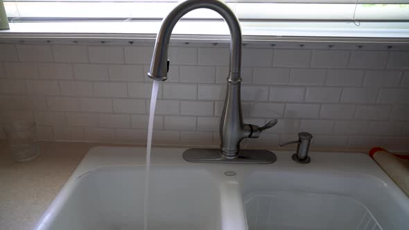 A wide shot as water runs from a stainless steel faucet into a kitchen sink in front of a tile backs
