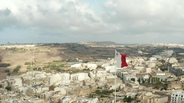 Flag of Malta Waving in Wind with View of City on Gozo Island, Aerial Close Up Slide Left