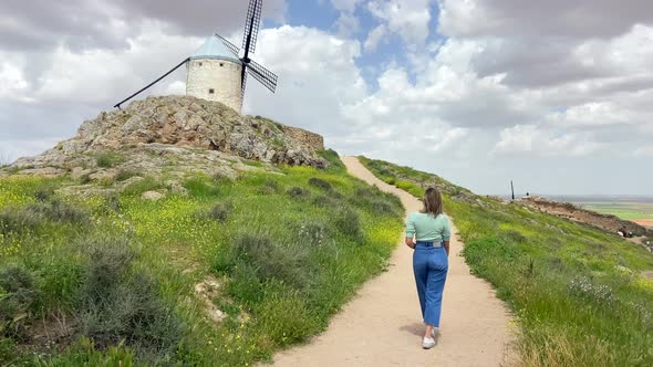 Young blonde country girl walking towards an old windmill in a meadow in sunny day