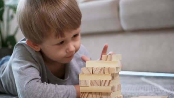 Child Builds a Wooden Tower for Playing Jenga Lying on the Floor at Home