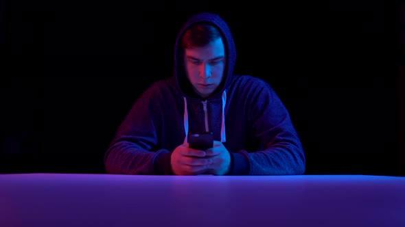 Young Man in a Hood with a Smartphone. Hacker Makes a Hack Through the Phone. Blue and Red Light