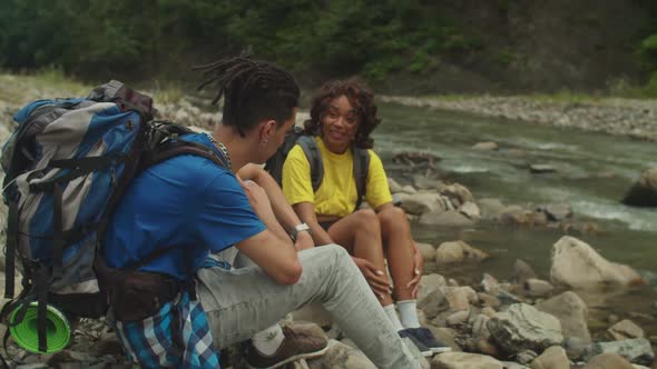 Positive Attractive Multiracial Couple Backpackers Relaxing on Mountain Riverbank on Hike