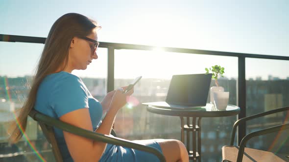 Woman Using Smartphone on the Balcony Against the Backdrop of the Sunset. Outdoor Home Office