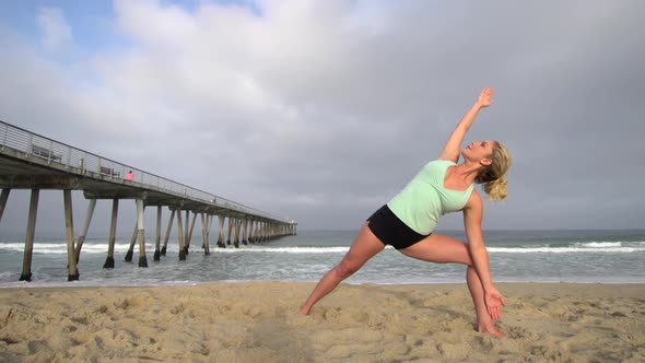 A young attractive woman doing yoga on the beach next to a pier.