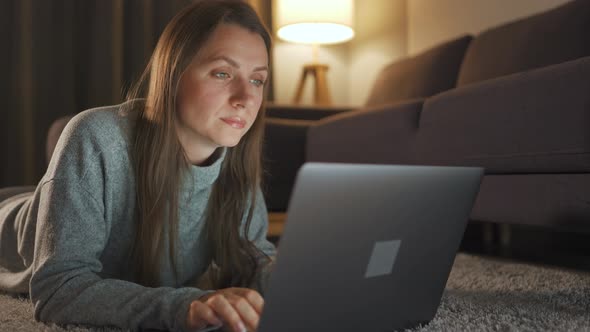 Woman Is Lying on the Carpet and Working on a Laptop in the Evening