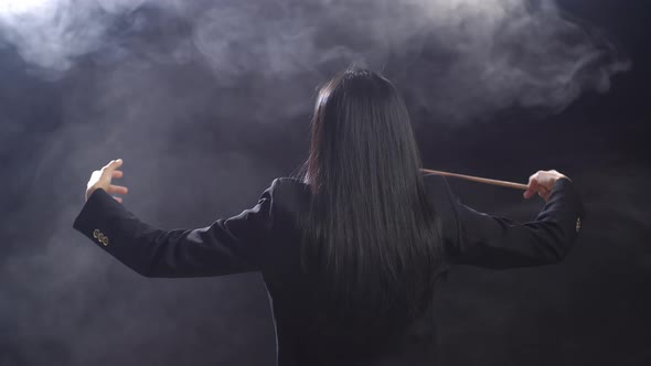 Back View Of Asian Conductor Woman Holding A Baton And Showing Gesture In The Black Studio With Fog