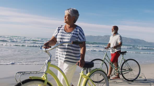 Smiling senior african american couple walking with bicycles at the beach