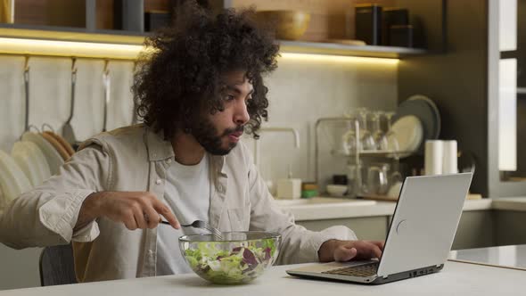 Arabian Man Eats Salad Working on Laptop at Table in Kitchen