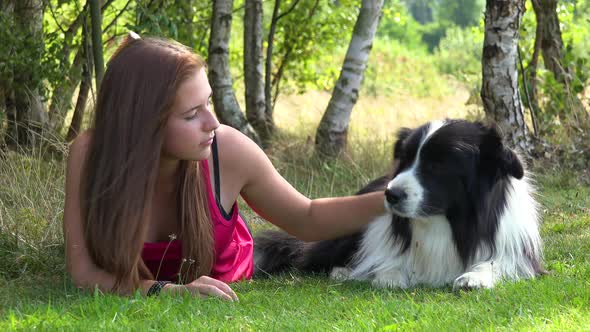 A Woman and a Border Collie Lie on Grass in a Meadow, the Woman Occasionally Pets the Dog and Looks