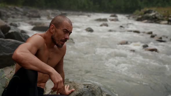 Muscular Man is Sitting on Stone Shore of Mountain River Admiring Landscape