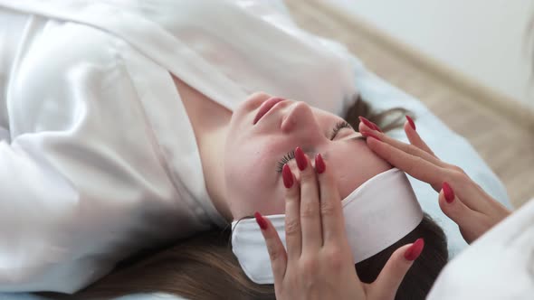 Hands of Beautician Making Massage on Face of Woman Lying with Closed Eyes on Couch