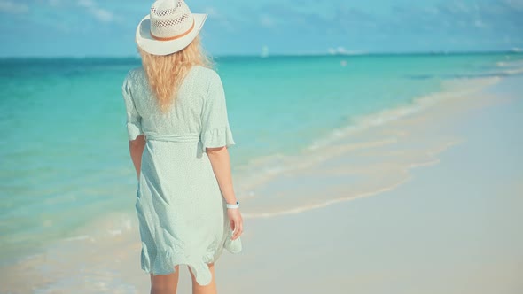 Woman In Hat Walks Along Beach On Caribbean Coast.Travel Mood Wind Blows Dress On Ocean Resort.