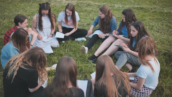 A Group of Female Students are Sitting in a Circle on a Meadow for Collective Work with Notebooks