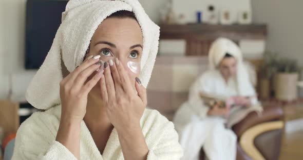 Pretty Young Caucasian Woman in White Bathrobe and Hair Towel Applying Eye Gel Patches