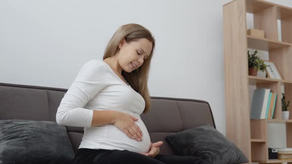 A Pregnant Fairskinned Woman is Sitting on a Cozy Sofa and Stroking Her Tummy