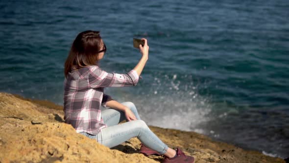 A Young Woman Is Sitting with a Phone on the Stones in Front of the Sea. Girl in Sunglasses Herself