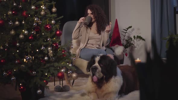 Beautiful Caucasian Woman Sitting on Armchair, Drinking Tea or Coffee and Eating Cookies. Her Lovely