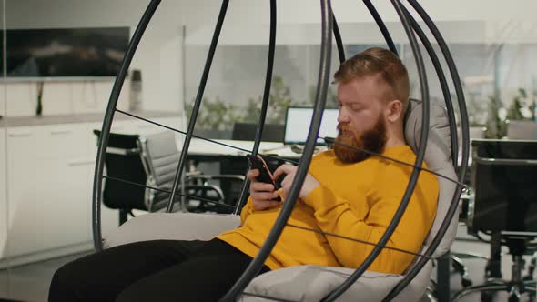 Bearded Guy Using Smartphone Sitting In Swing Chair In Office