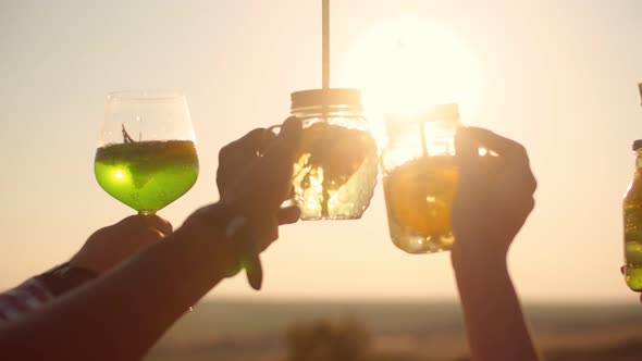 Hands holding high four glasses with cocktails against background of bright sun, closeup. Friends