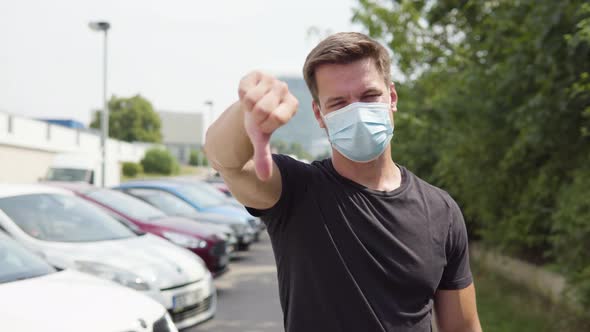 A Young Man in a Face Mask Shows a Thumb Down To the Camera and Shakes His Head in a Parking Lot