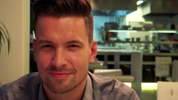 A Young, Handsome Man Sits at a Table in a Restaurant and Smiles at the Camera - Closeup