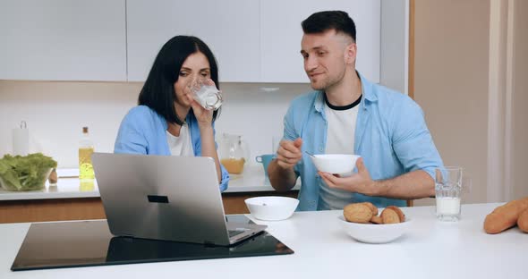 Couple Sitting in Front of Computer Screen at the Kitchen Table and Having Breakfast