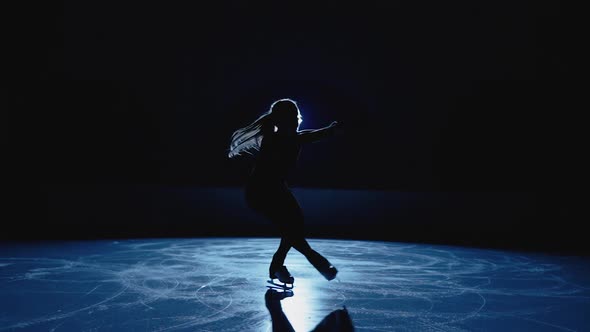 Young Figure Skating Artist Rotates on Ice Arena in Dark Against Backdrop of Bright Blue Spotlight