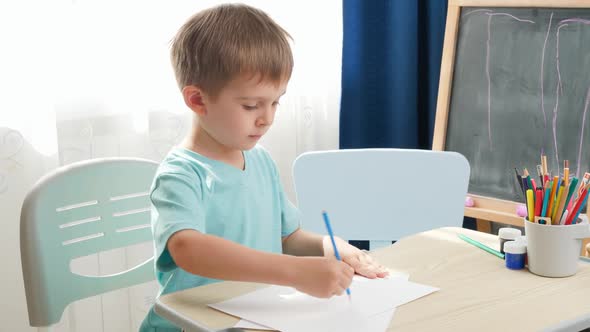 Concentrated Little Boy Drawing or Writing with Pencil While Sitting Behind School Desk at Home or